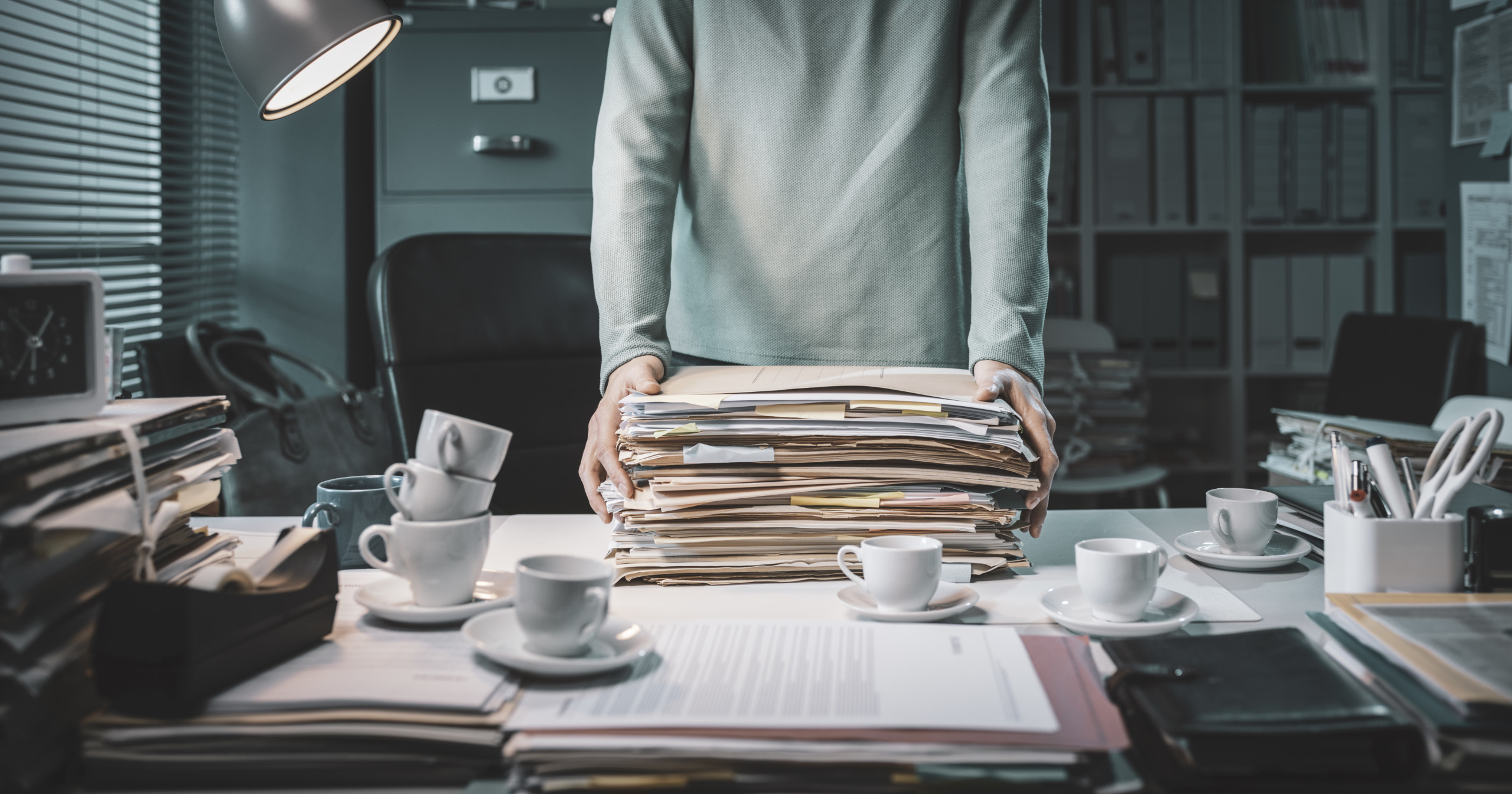 Male standing behind a desk, from shoulders to waist. He has his hands on a large stack of files which are on the desk, as are 6 coffee cups and more paper files.